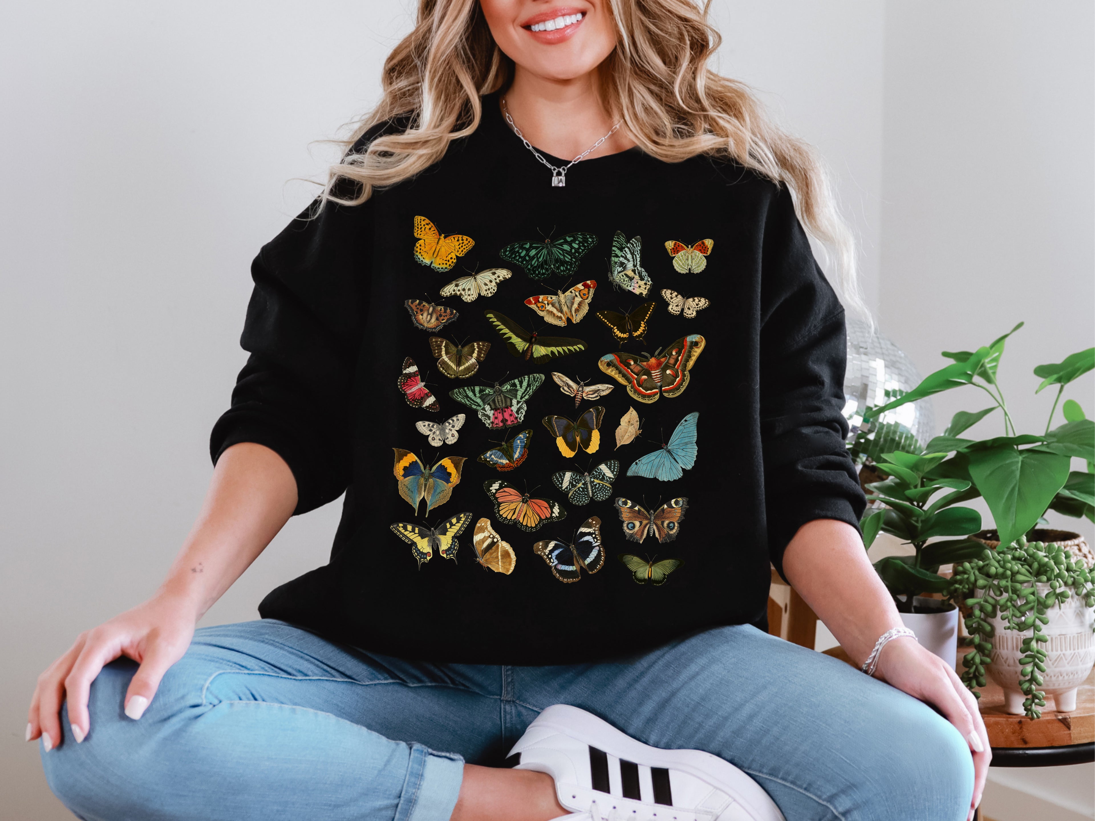 Woman wearing a black Gildan crewneck sweatshirt featuring a colorful butterfly and moth design, sitting casually with jeans and sneakers.