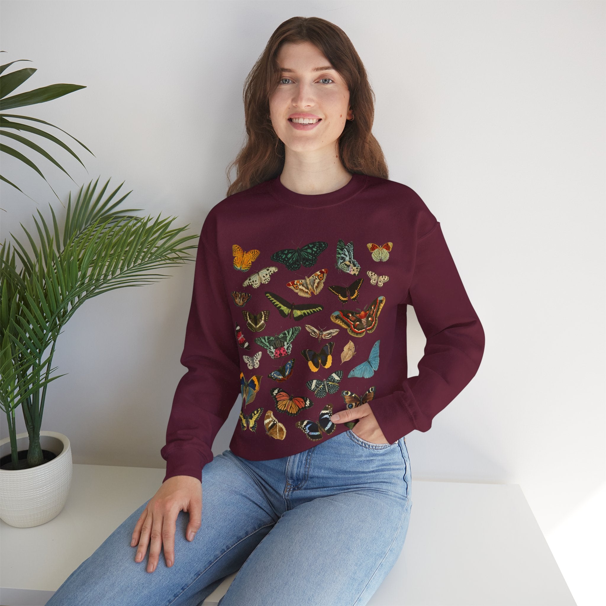 Woman wearing a maroon sweater with butterfly designs sitting on a white surface with a plant in the background.