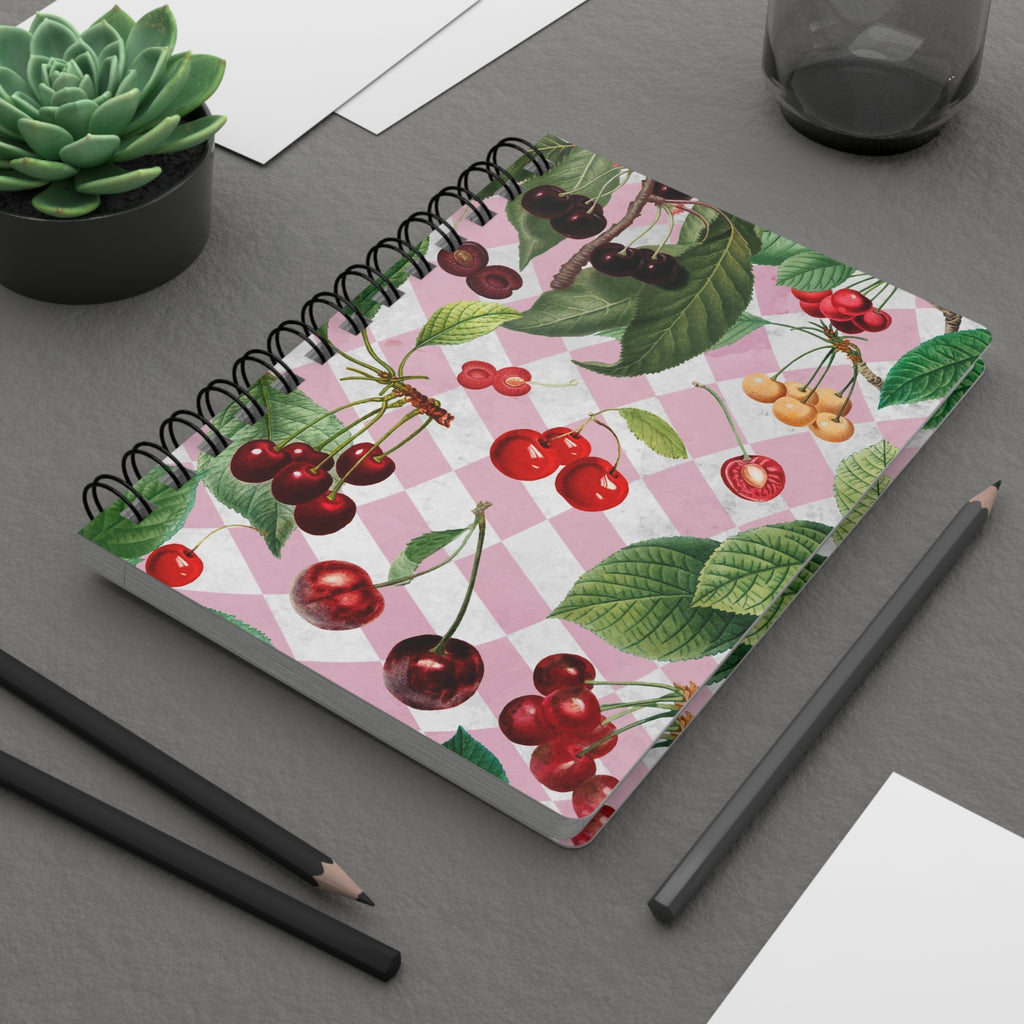 Notebook with cherry design on a gray surface with pencils and a plant.
