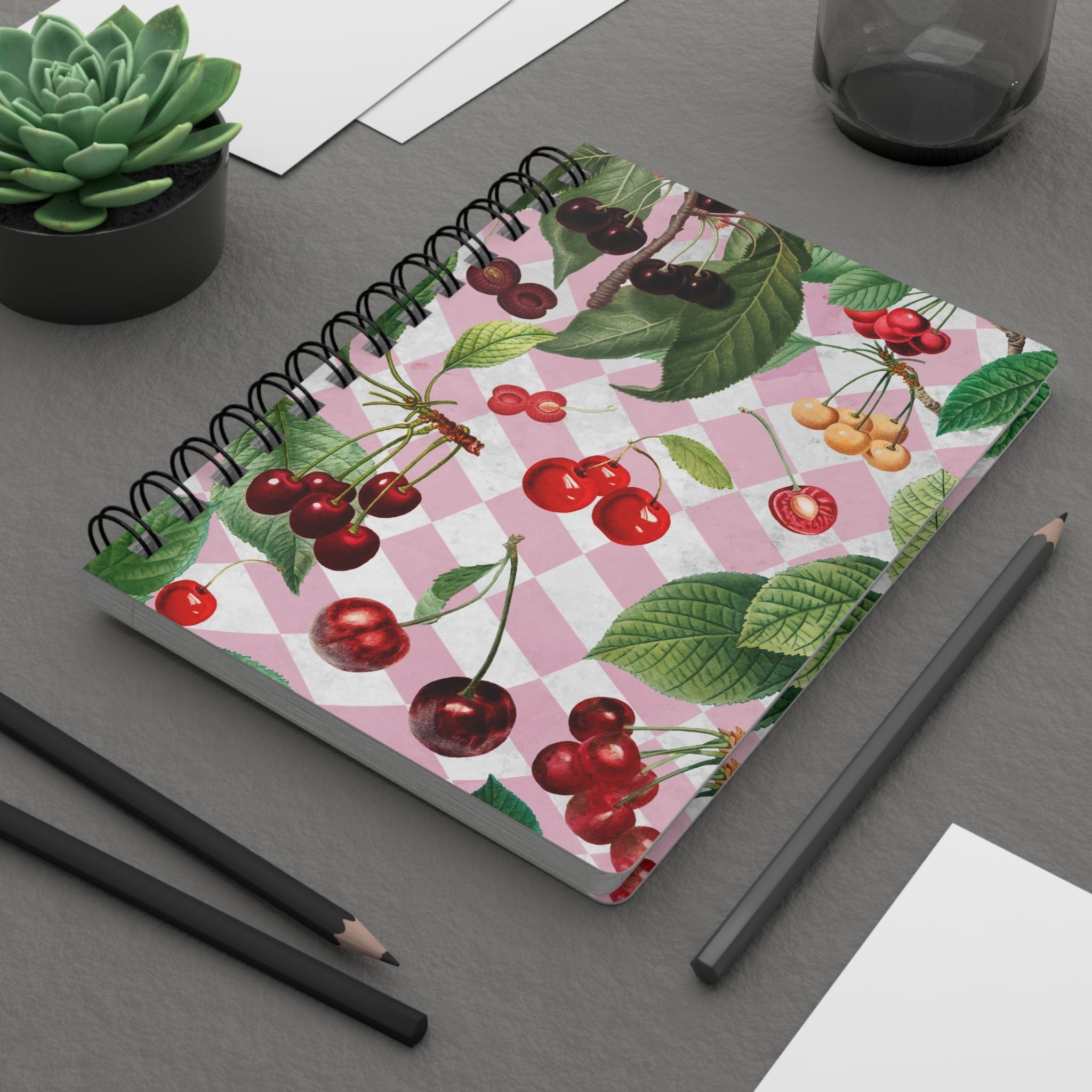 Notebook with cherry design on a gray surface with pencils and a plant.
