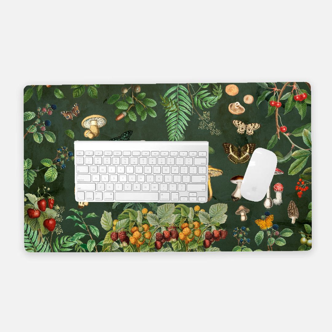 “Cottagecore mushroom desk mat with vintage botanical artwork featuring forest plants, berries, butterflies, and mushrooms on a deep green background. Keyboard and mouse to give reference to sizing.