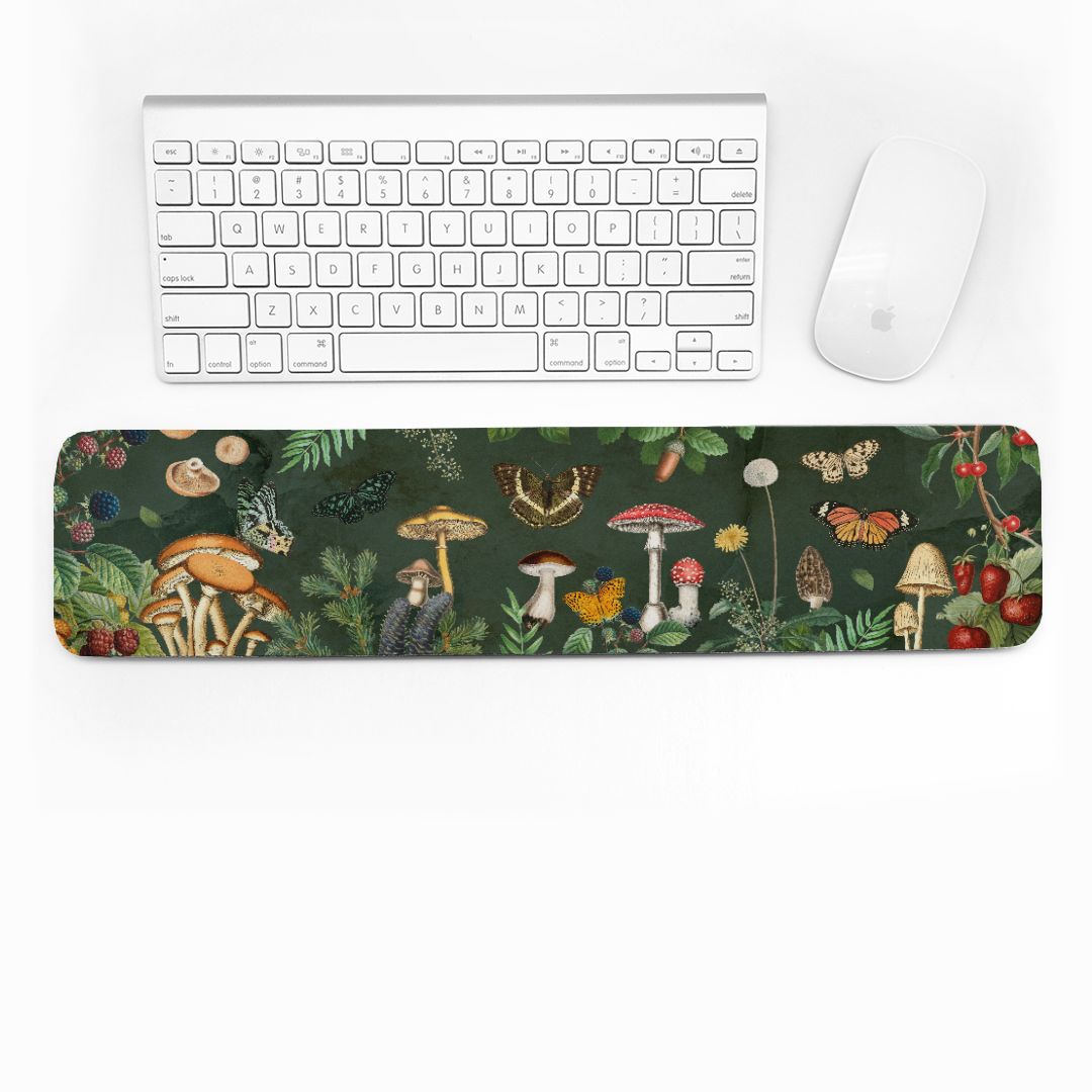 Desk setup with a forest-themed cottagecore keyboard wrist rest, keyboard, and mouse on a white surface. Mushrooms, butterflies, and greenery on a dark green background.