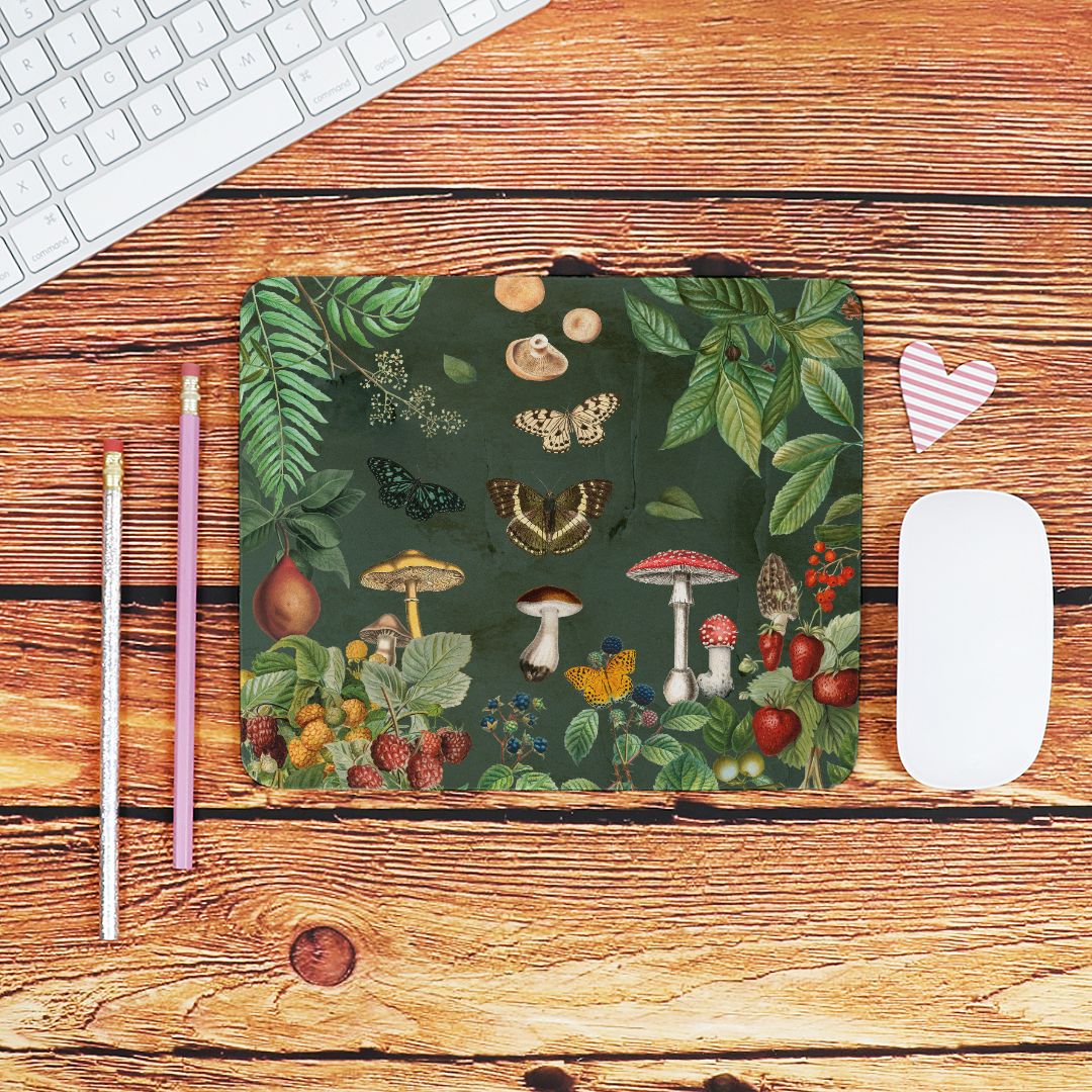 Desk setup with a nature-themed mouse pad including butterflies berries, mushrooms & greenery, keyboard, pencils, and heart-shaped object on a wooden surface.