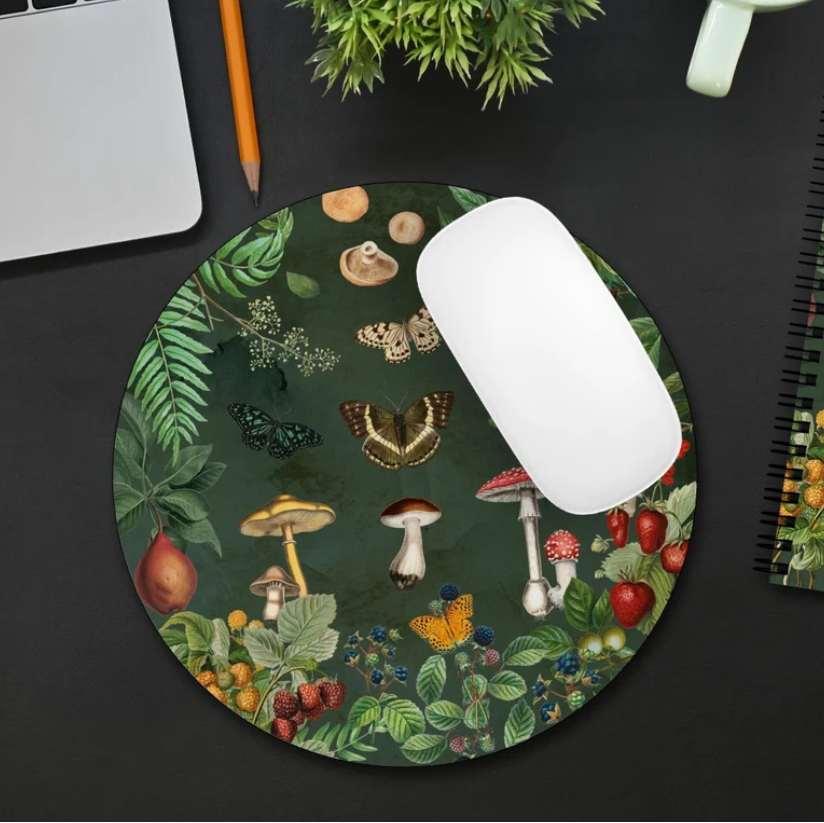 Desk setup with a round cottagecore mouse pad, notebook, and computer mouse on a dark surface. Mushroom, butterfly, berries and greenery print.