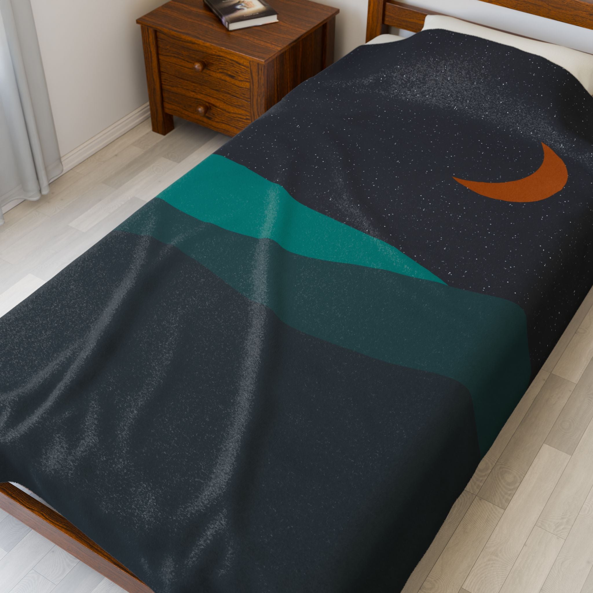 Bed with a dark blanket featuring a crescent moon design in a bedroom setting. Featuring a dark navy night sky with an orange crescent moon with teal colored desert hills.