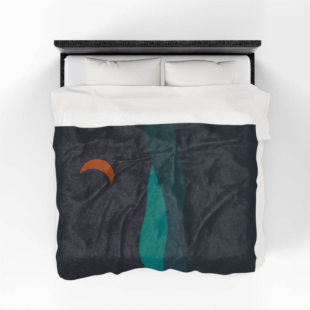 Bed with a white comforter featuring a throw blanket on top with a teal and navy and a crescent moon design on a white background.
