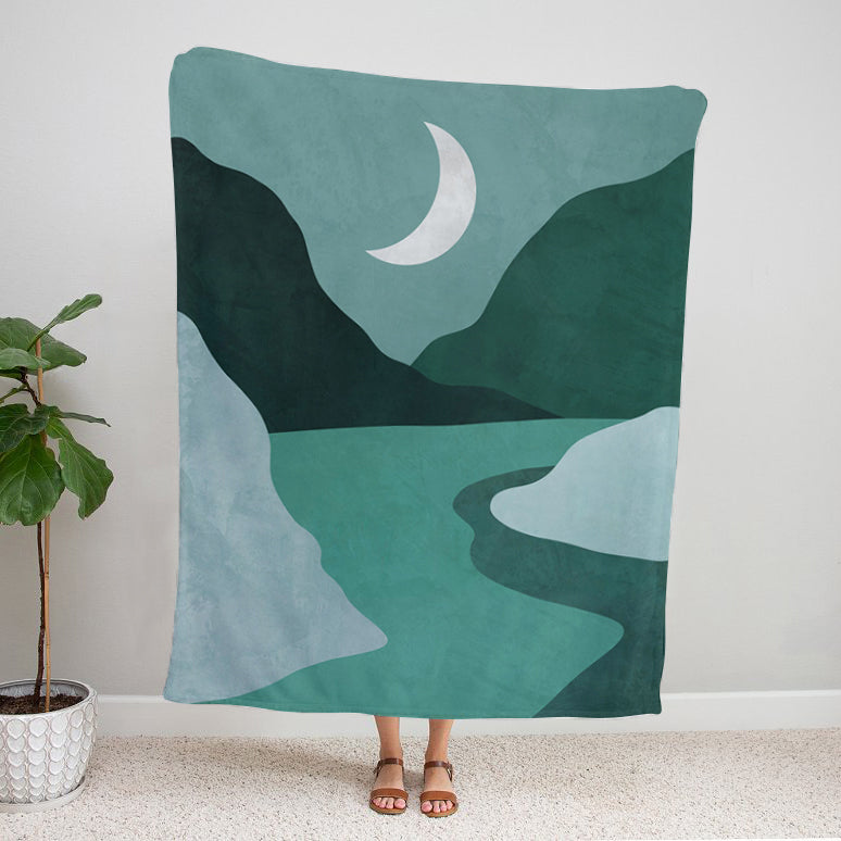 Person holding a Soft velveteen blanket with moon and mountain landscape design in blue and green tones against a plain wall. 50x60 inch blanket