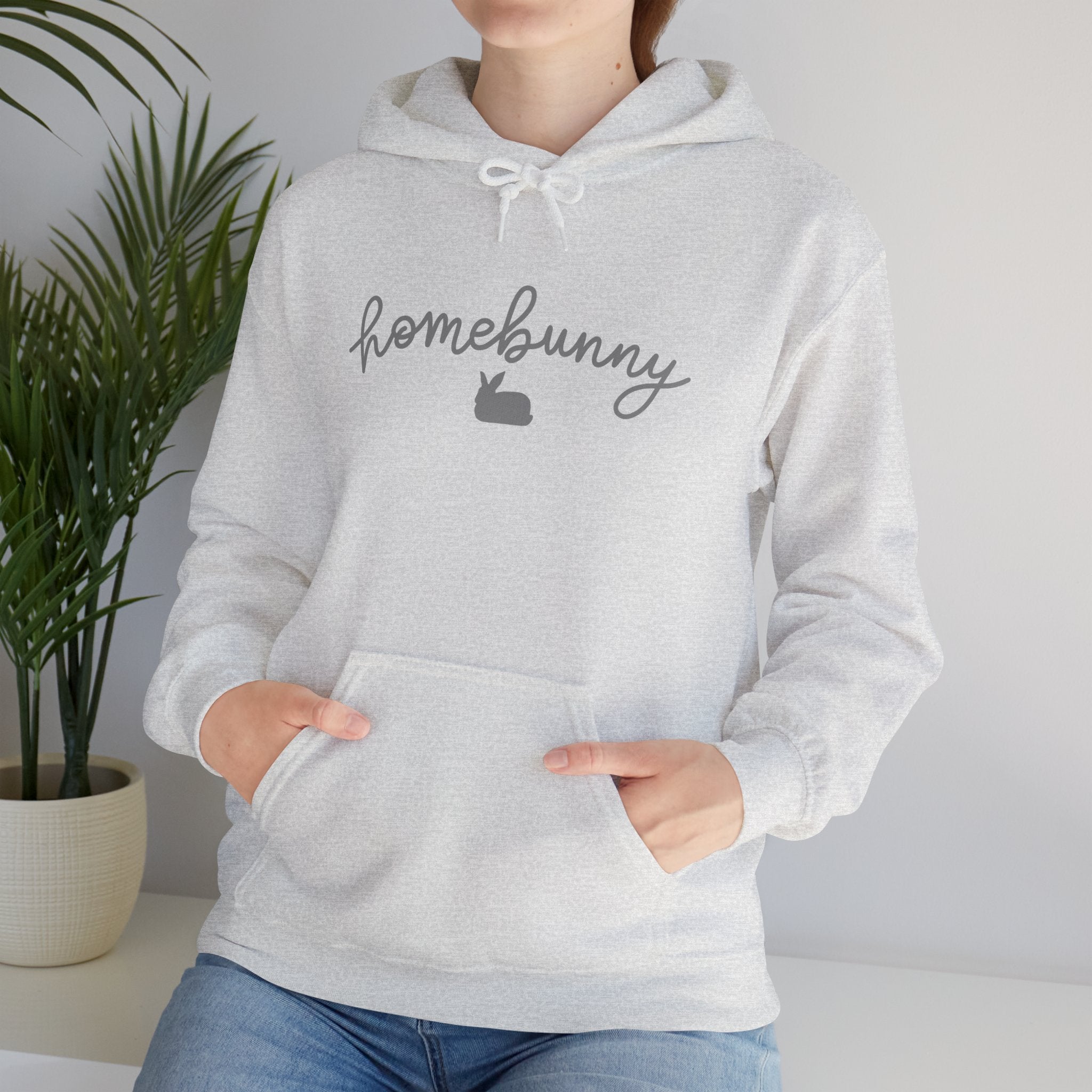 Person wearing a ash gray hoodie with 'homebunny' text, standing indoors next to a plant.