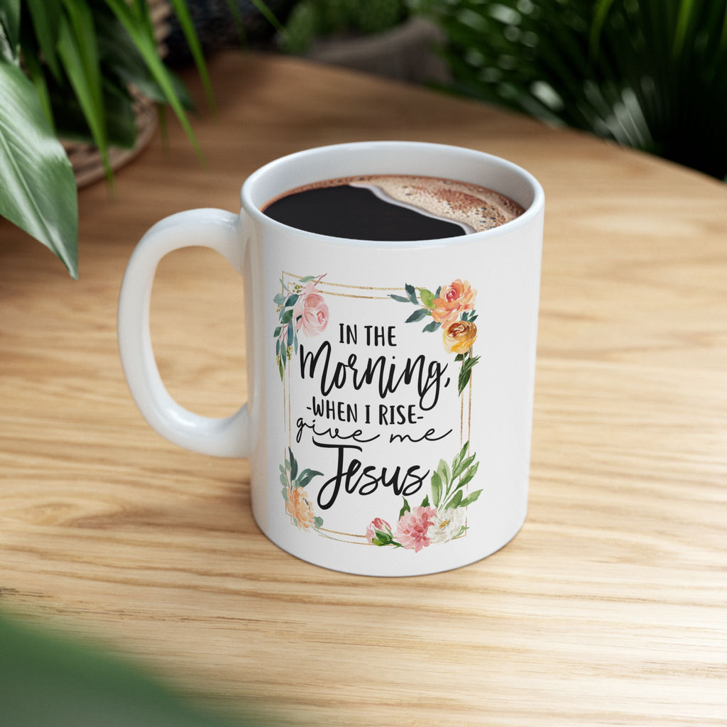 White ceramic coffee mug filled with coffee, featuring floral artwork and the quote “In the Morning When I Rise Give Me Jesus,” sitting on a wooden table with green plants in the background.