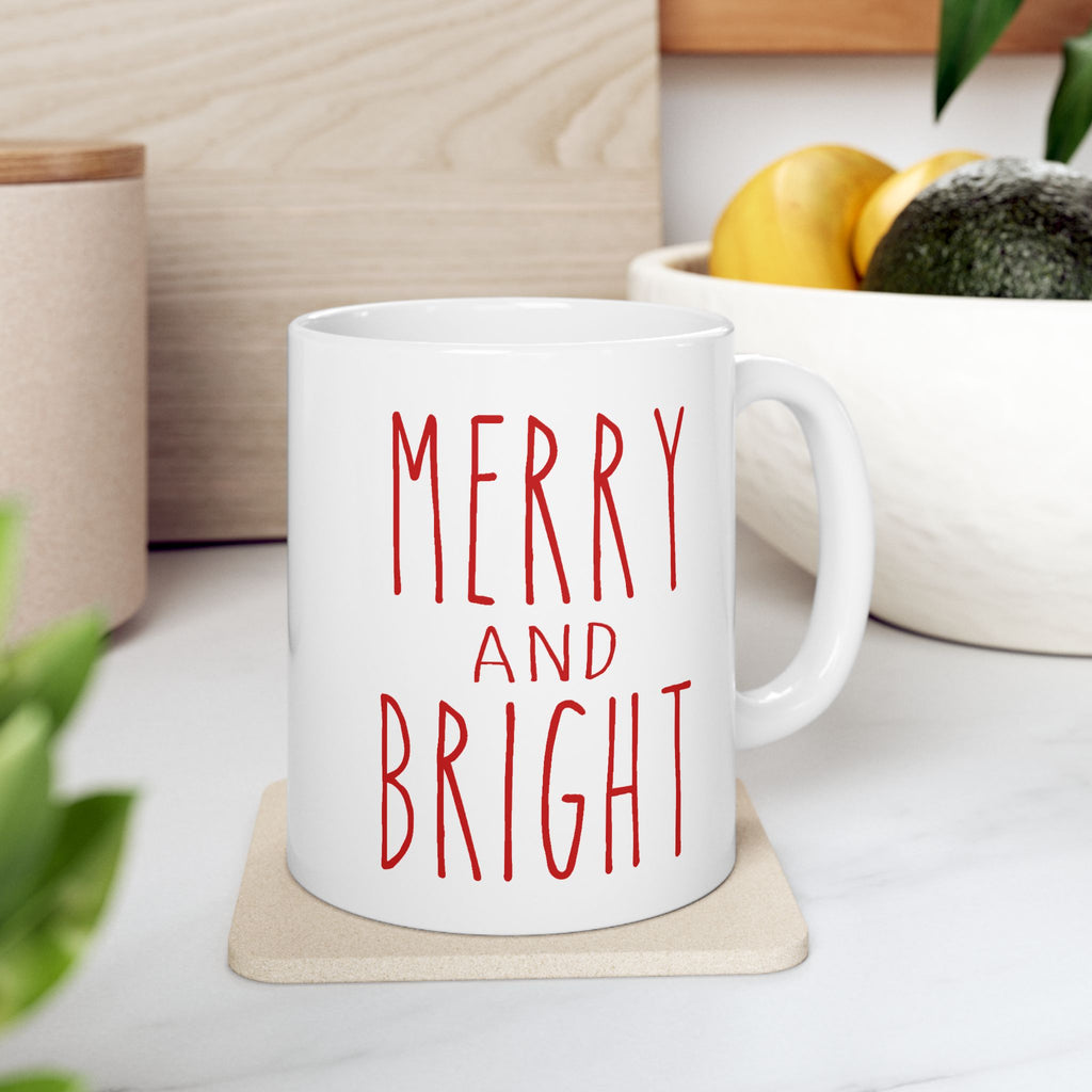 White mug with 'Merry and Bright' text on a coaster, surrounded by plants and a bowl of fruit.