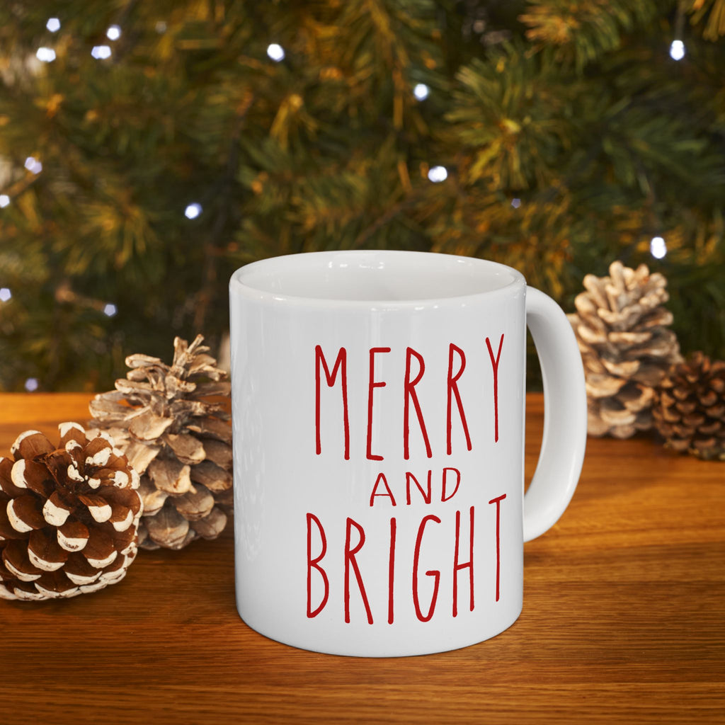 White mug with 'Merry and Bright' text in red, placed on a wooden surface with pinecones and a Christmas tree in the background.