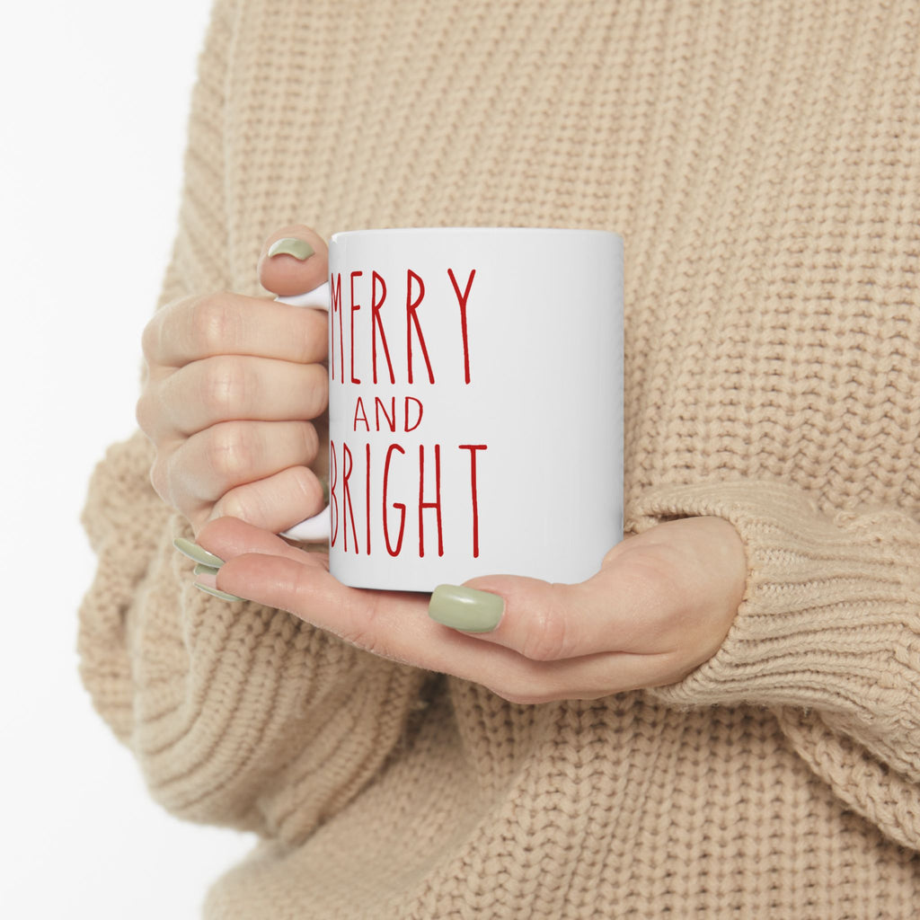 Person holding a white mug with 'Merry and Bright' text, wearing a beige sweater.