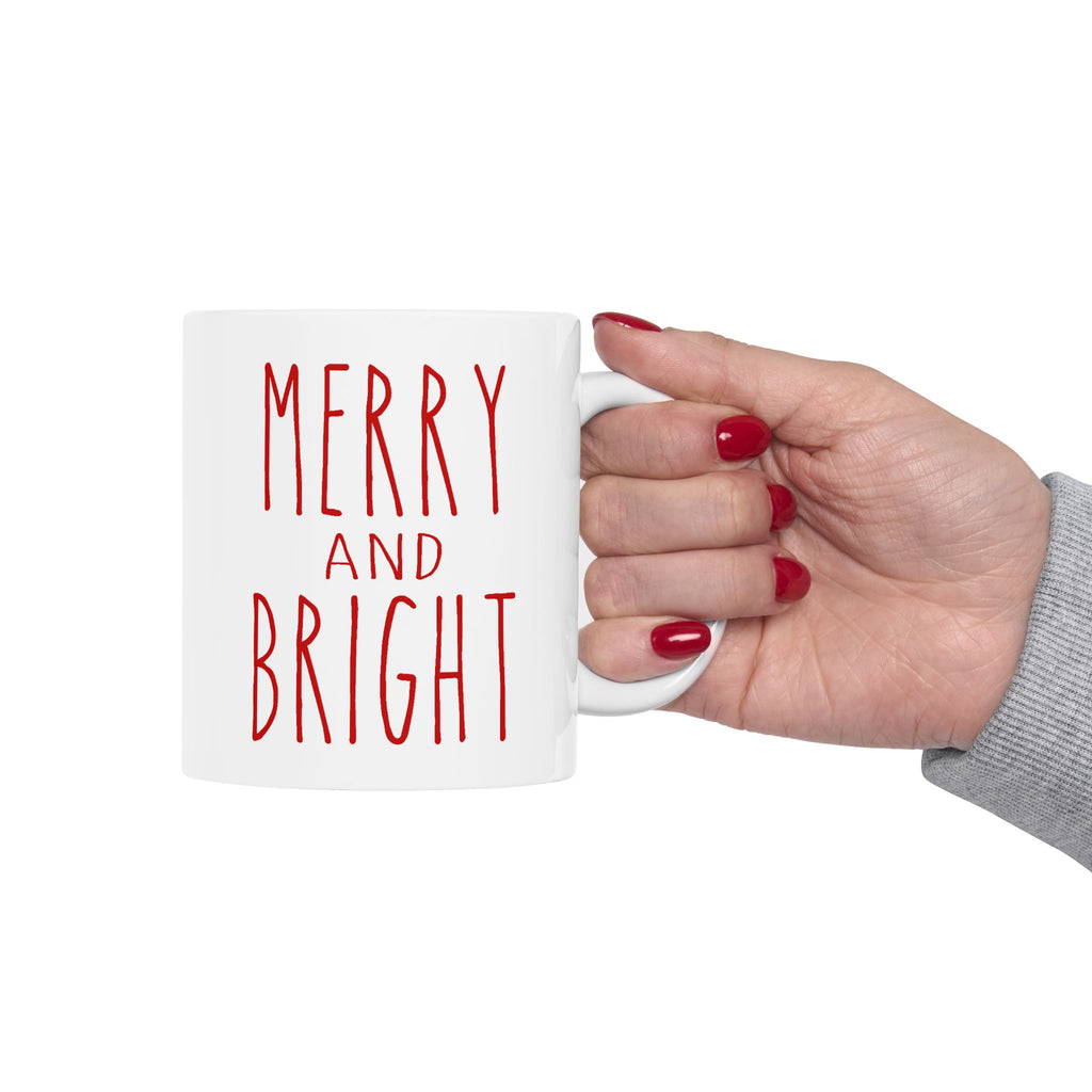 White mug with 'MERRY AND BRIGHT' text held by a hand with red nail polish on a white background