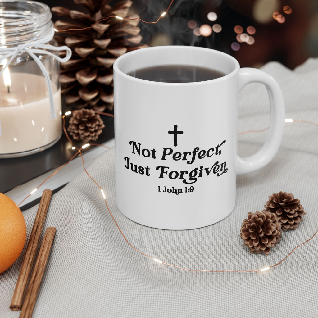 White mug with 'Not Perfect, Just Forgiven' text on a table with candles and pinecones.