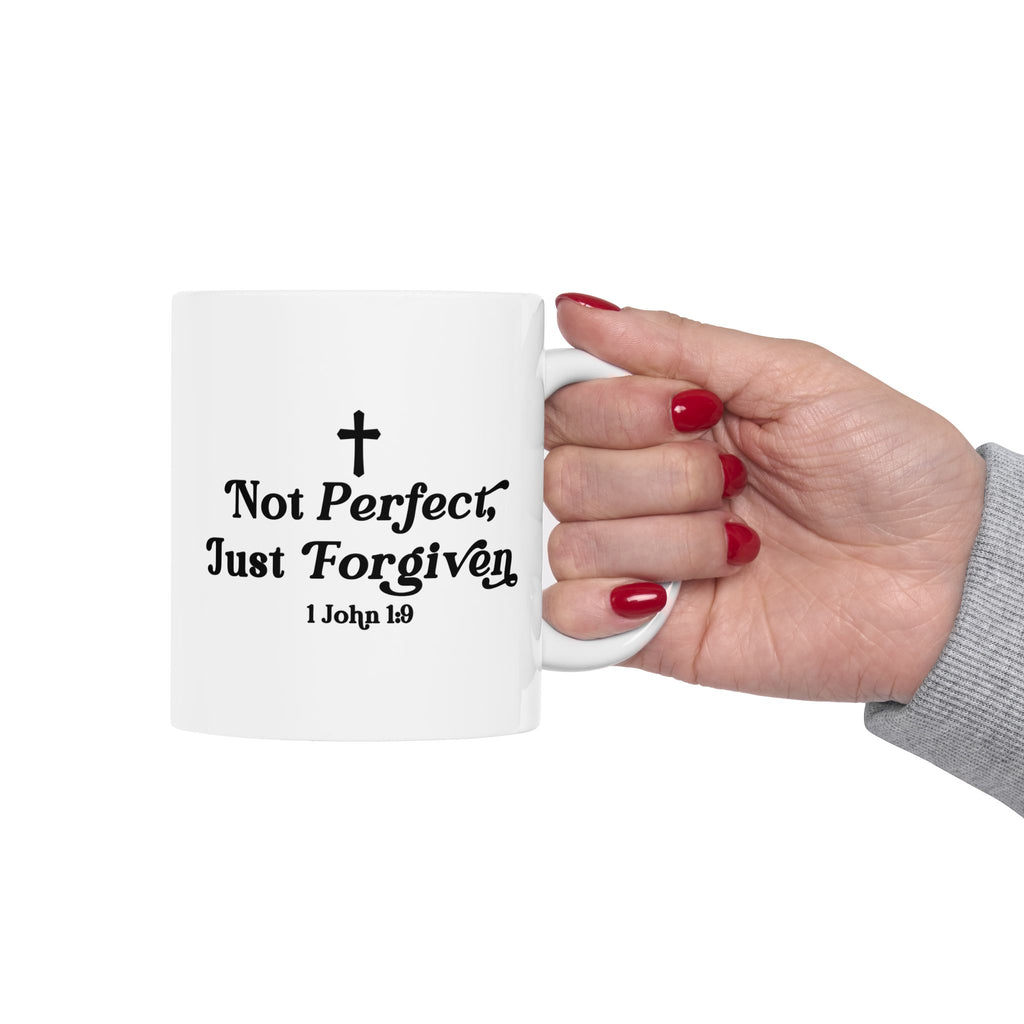 White mug with 'Not Perfect, Just Forgiven' 1 John 1:9 text held by a hand with red nail polish on a white background