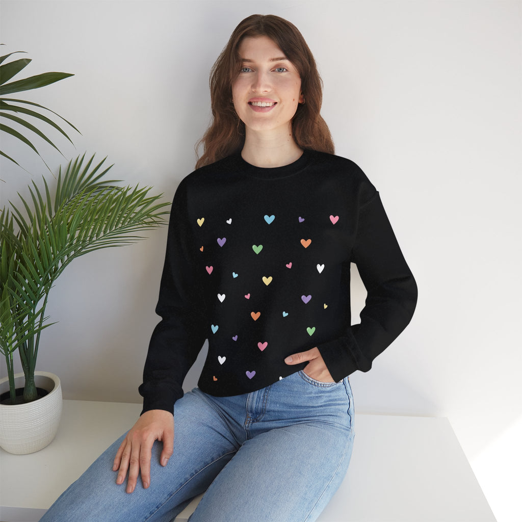 Woman wearing a black sweater with colorful heart patterns sitting on a white surface with a plant in the background.