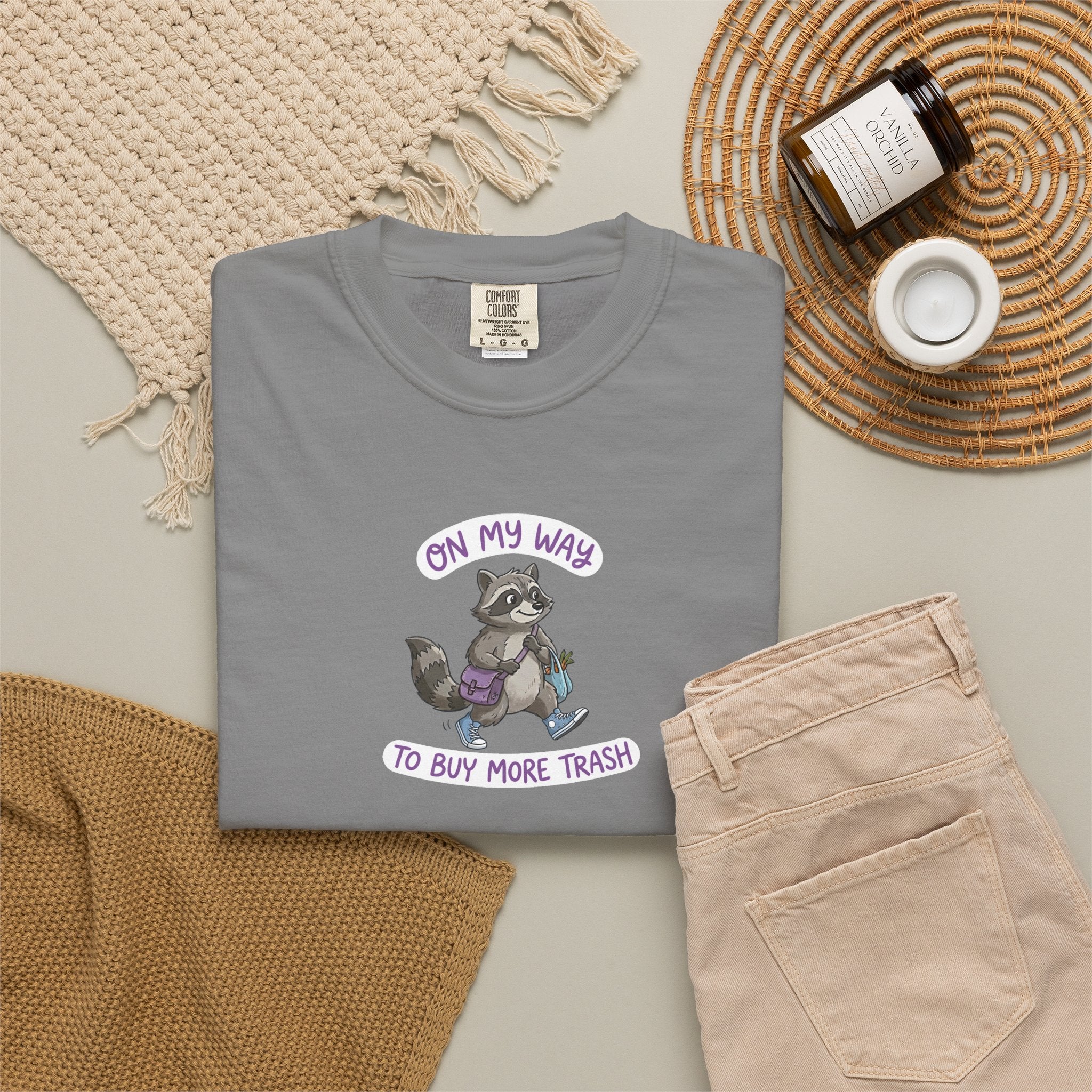 Gray t-shirt with a raccoon graphic and text on a neutral surface with accessories.
