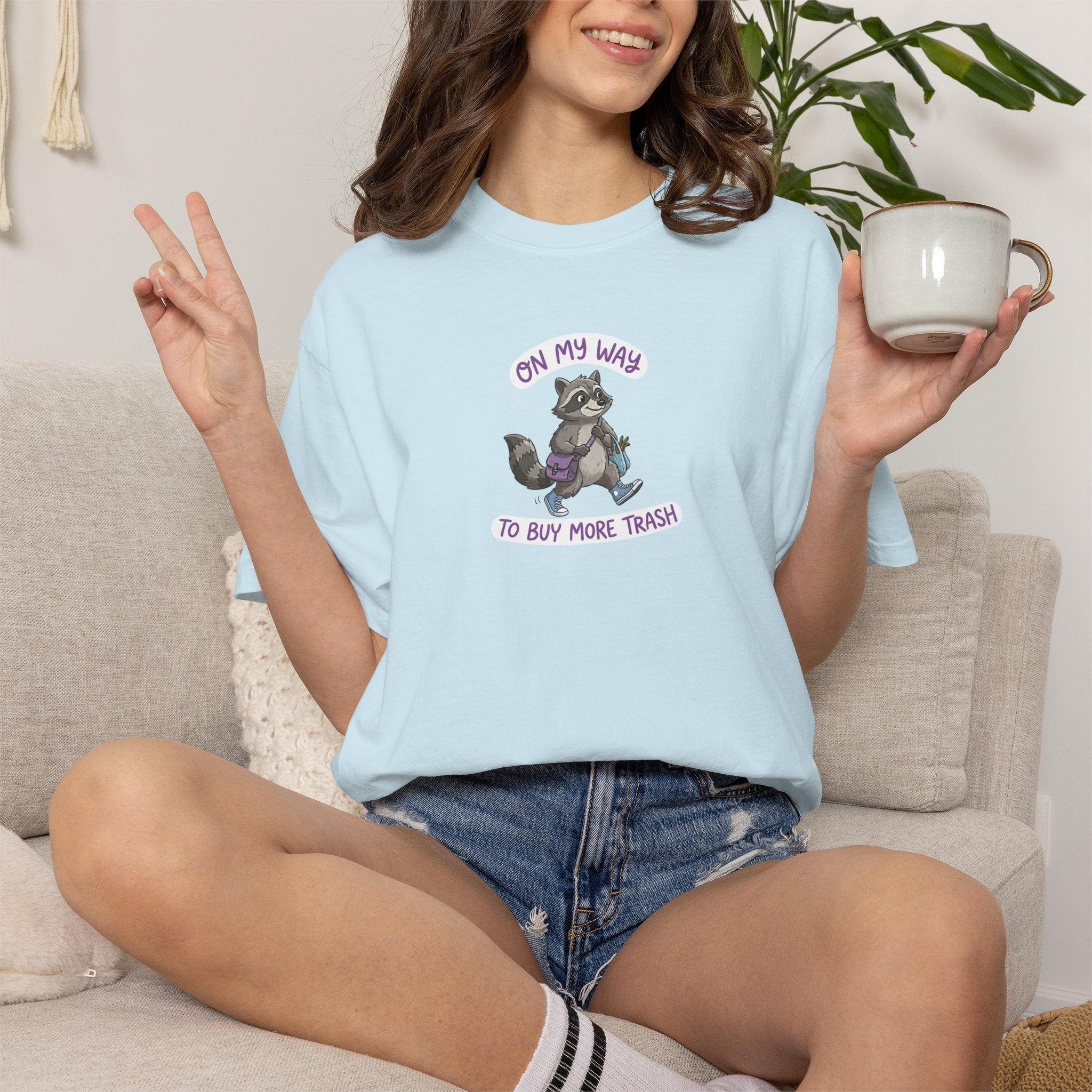 Person wearing a light blue t-shirt with a raccoon graphic and text "On My Way To Buy More Trash", sitting on a couch holding a mug.