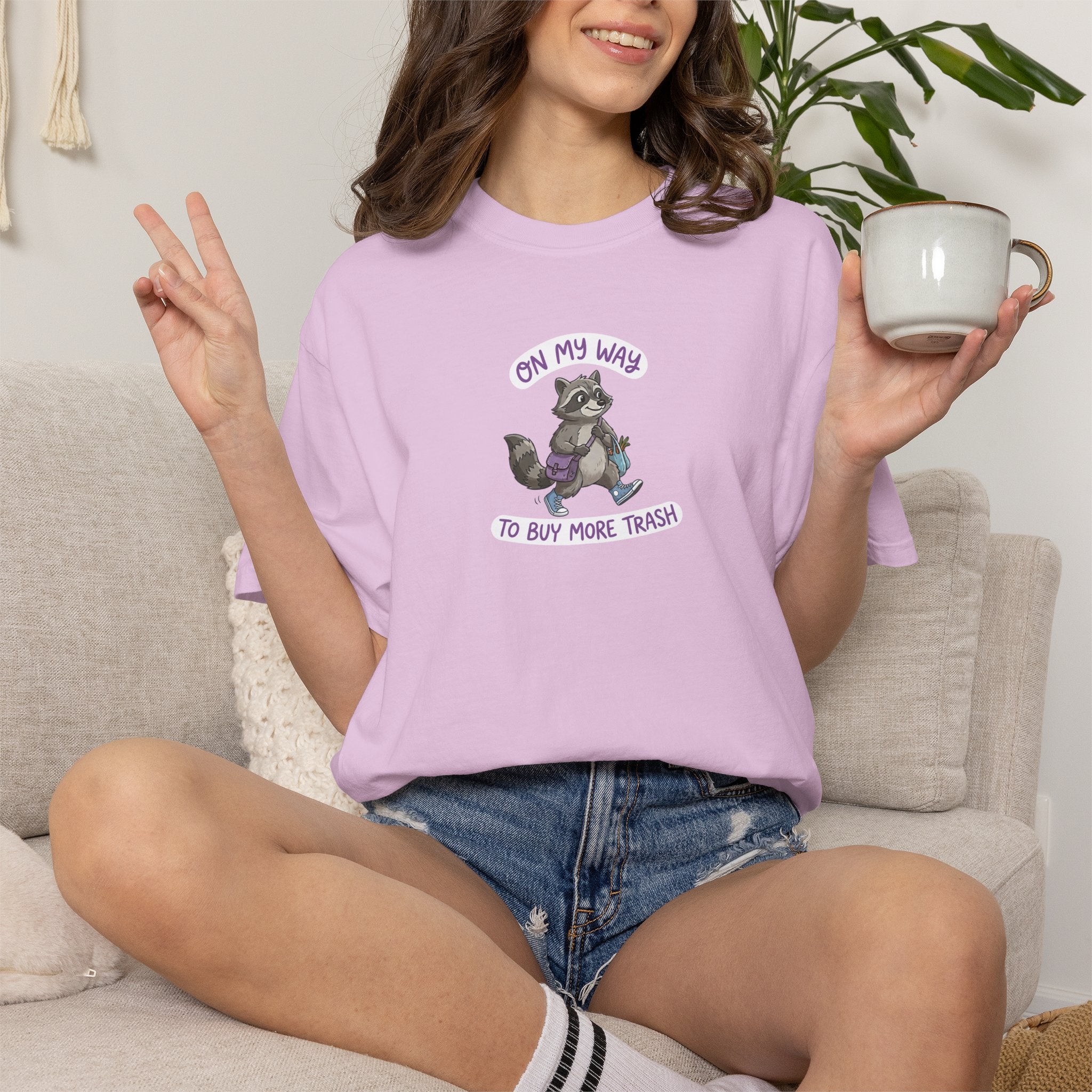 Person wearing a light pink t-shirt with a raccoon graphic and text, sitting on a couch holding a mug.